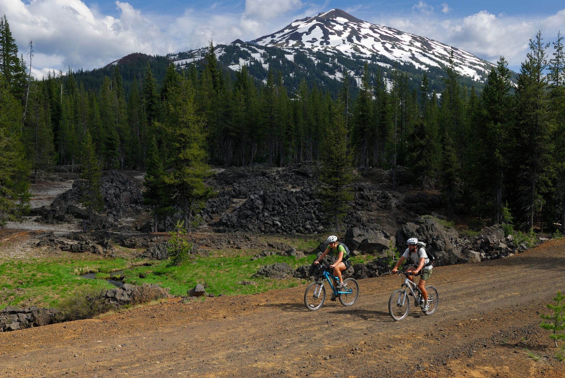 Cyclists riding on a dirt trail with a snow-capped mountain and pine forest in the background
