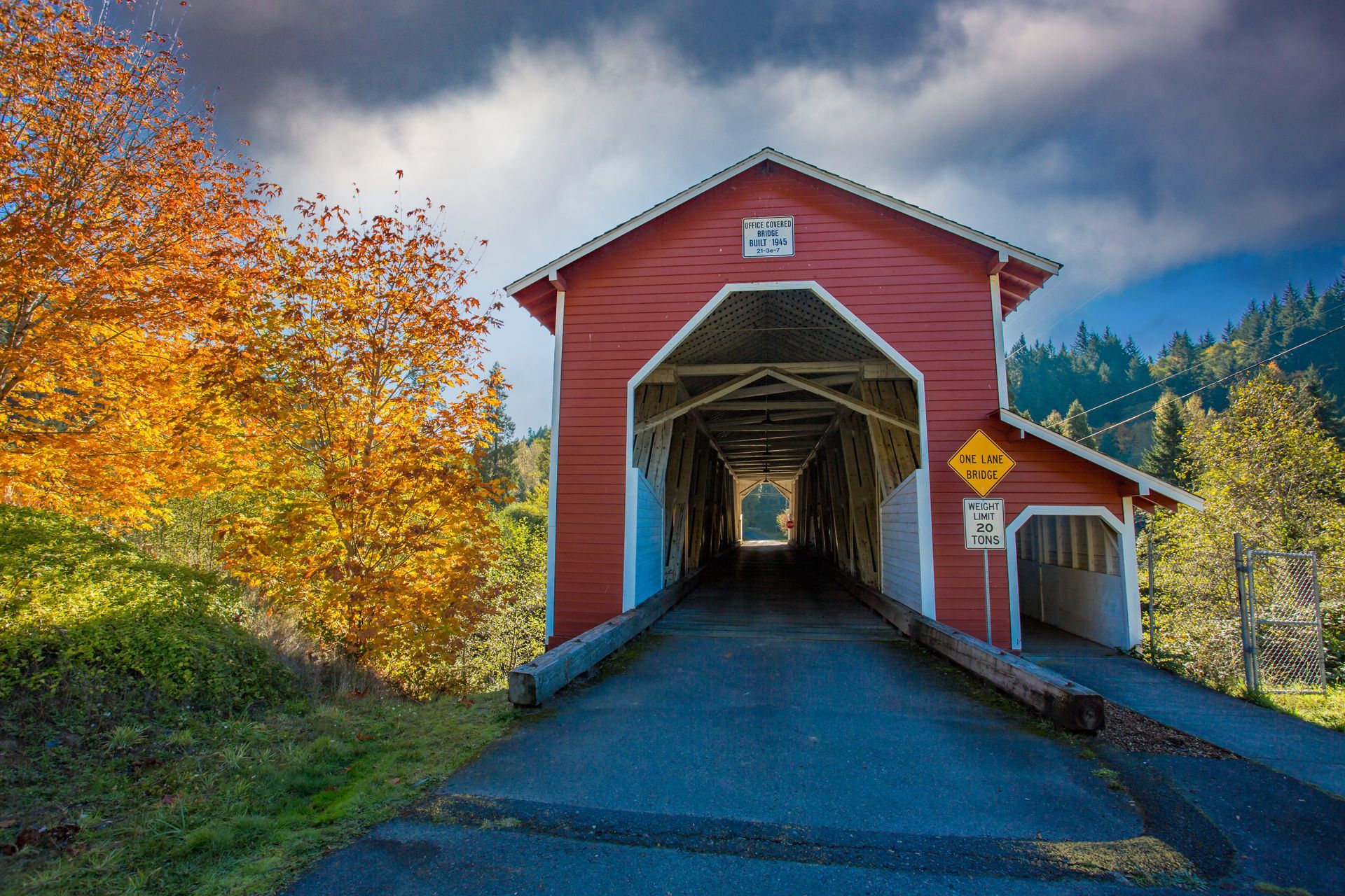 Red covered bridge on a road, framed by autumn trees and mountains under a cloudy sky