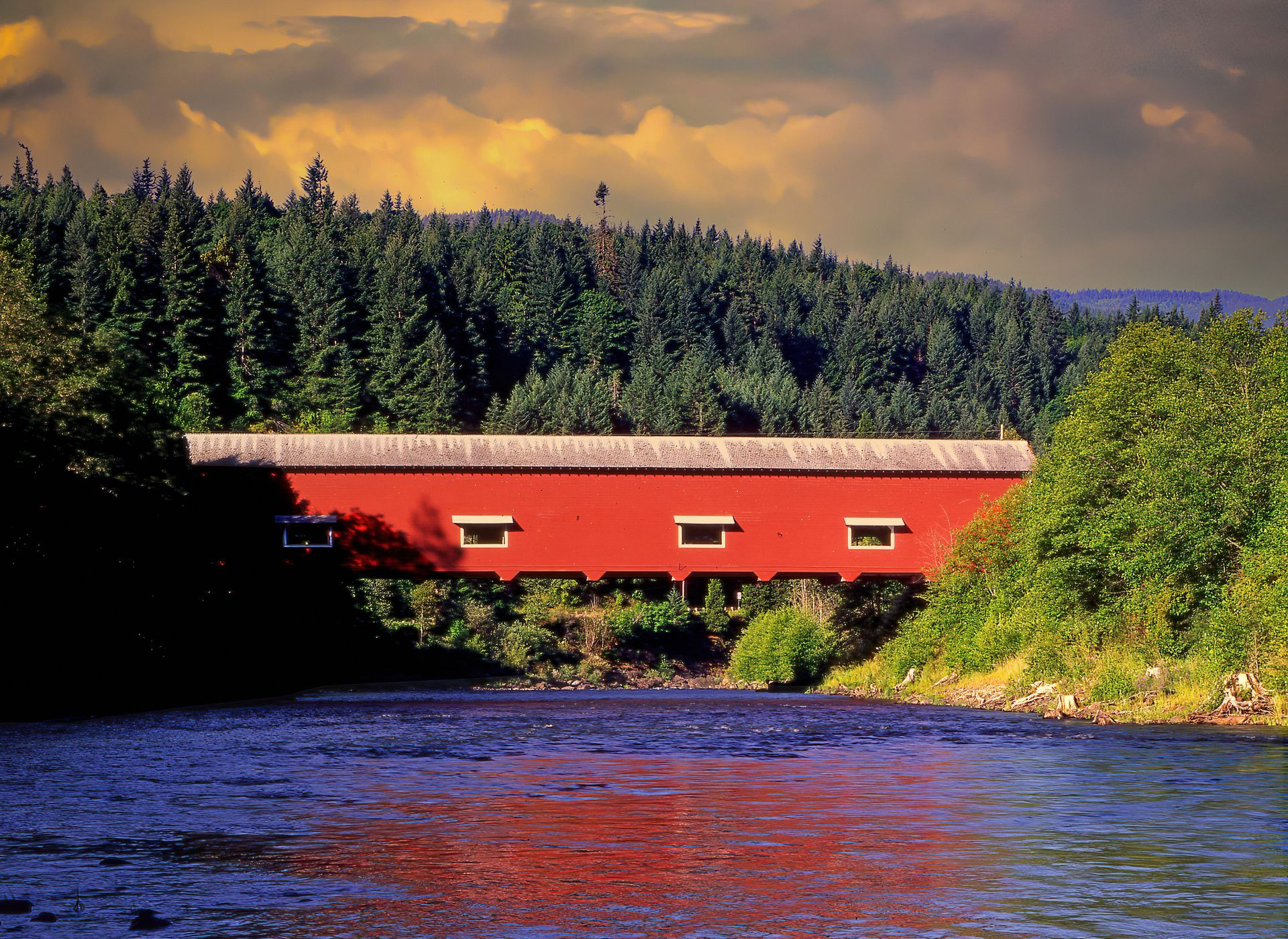 Red covered bridge over a river, surrounded by dense green trees under a warm sunset sky