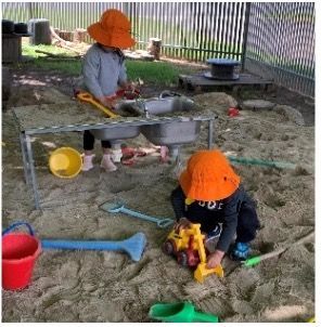 Two children in orange hats playing in a sandbox with toys and a sink.
