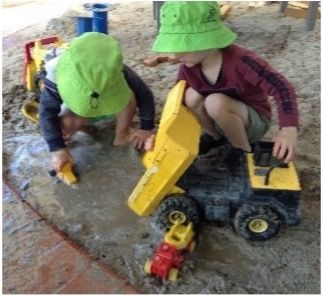 Two children in green hats playing with toy trucks in a sandbox.