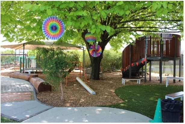 Playground with tree, play structure, decorations, wood chips, and grass.