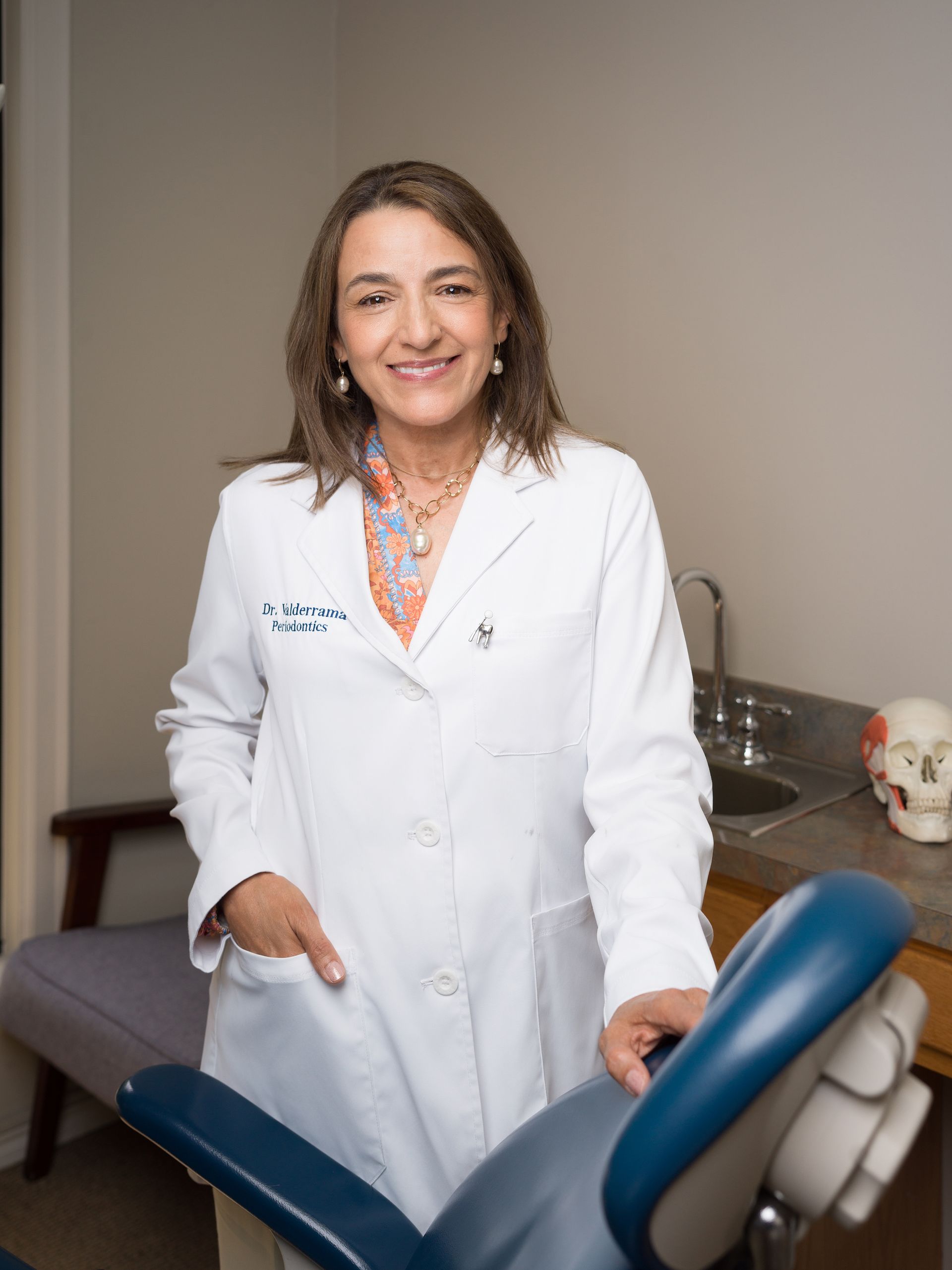 Smiling doctor in a white coat standing in a medical exam room beside an exam chair.