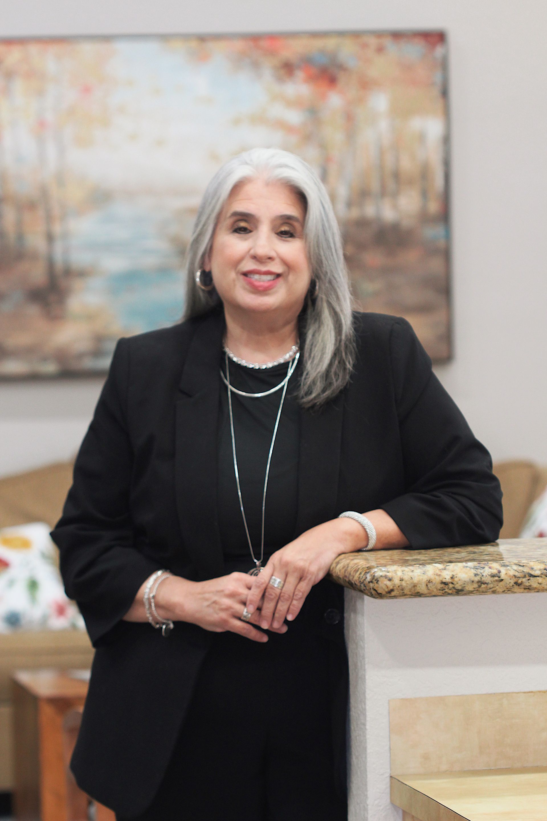 Woman in a black suit leaning on a marble counter, smiling in a bright room with abstract wall art.