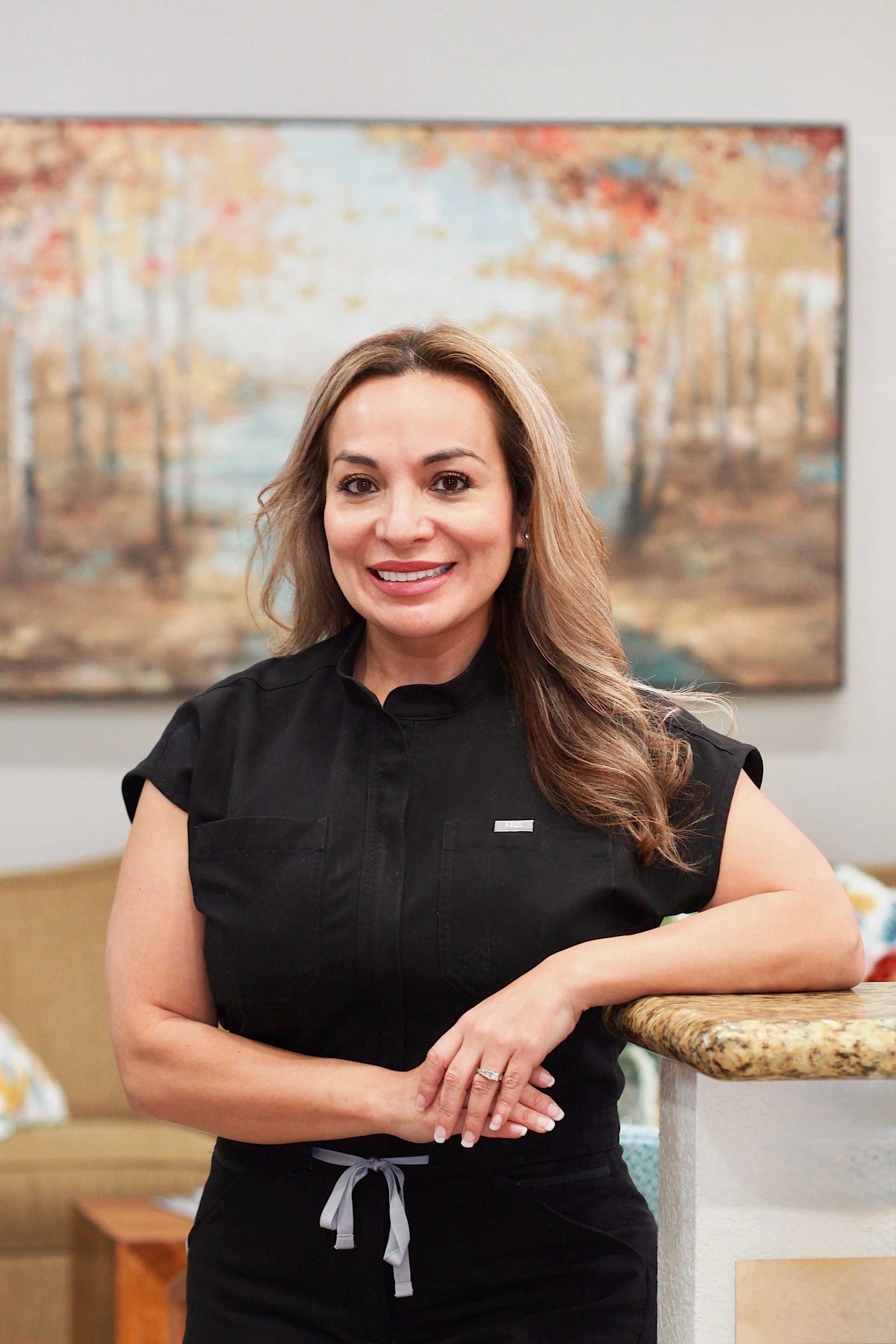 Woman in a black top smiling by a counter, with a framed autumn landscape painting behind her.