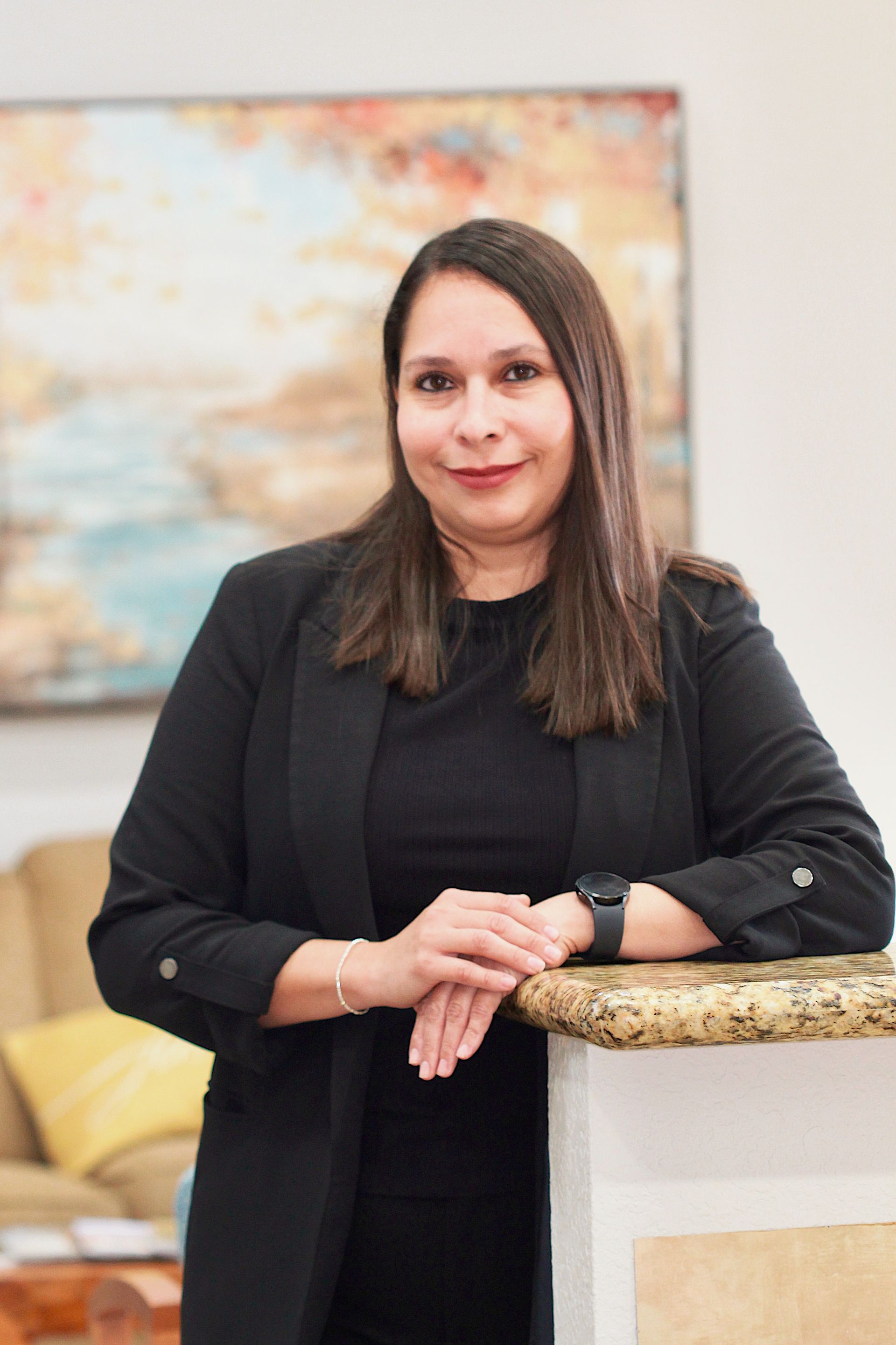 Woman in a black blazer leaning on a granite counter in a bright office, with framed artwork behind her.