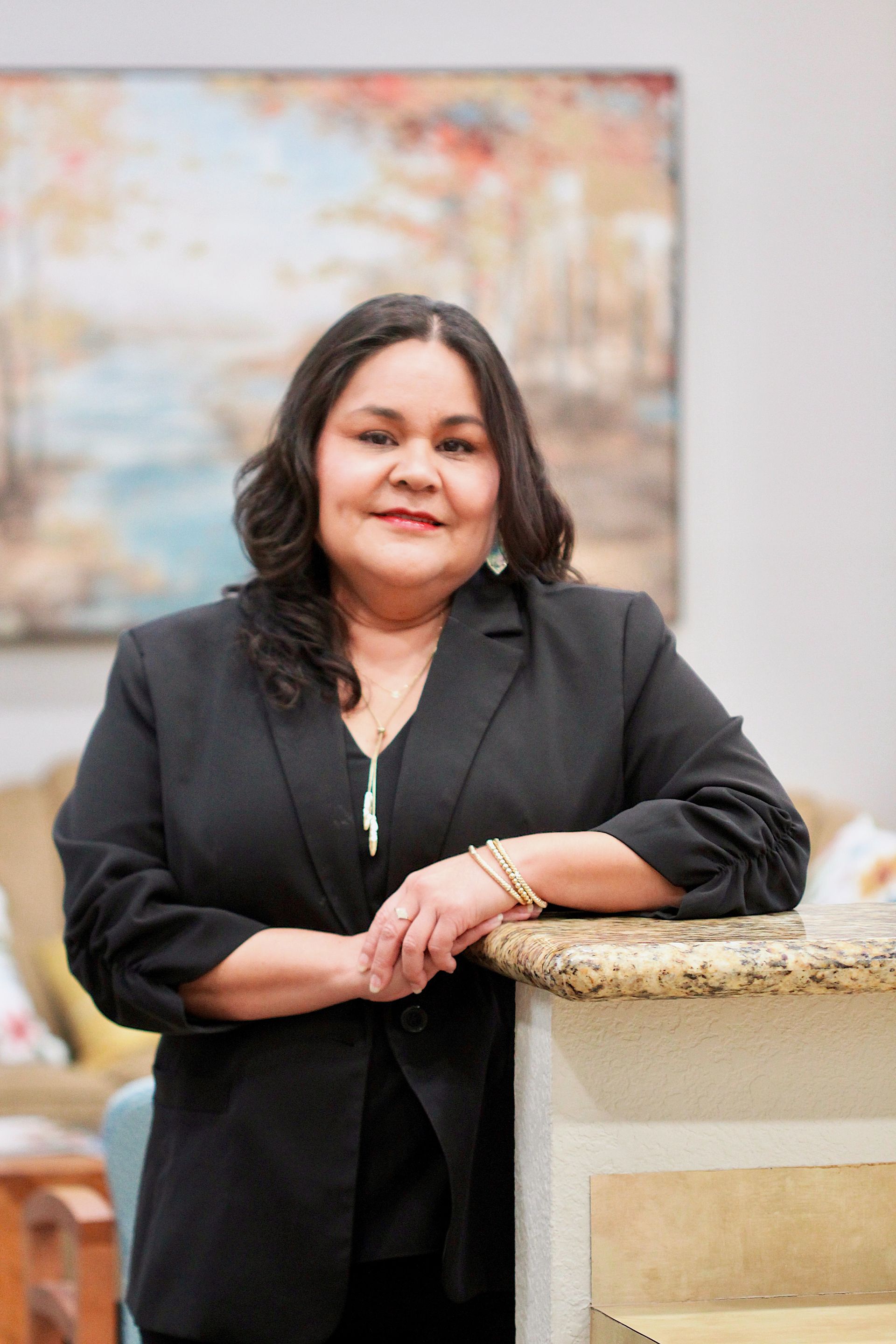 Woman in a black blazer leaning on a stone counter, smiling in a softly blurred indoor setting.