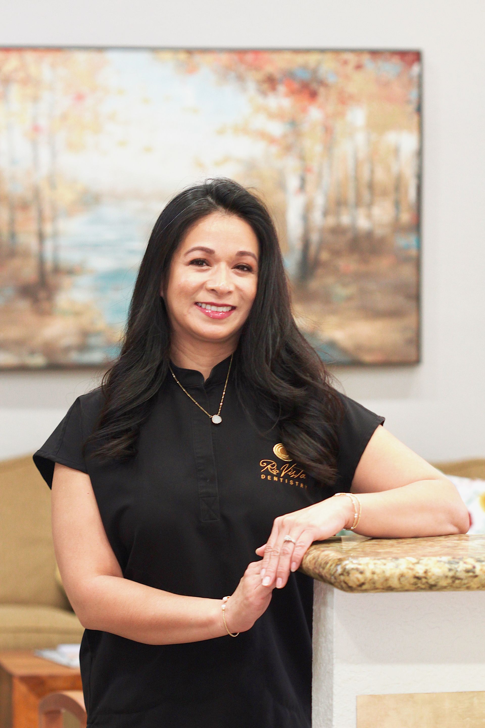 Woman standing by a marble counter in a black top, smiling in front of a framed landscape painting.