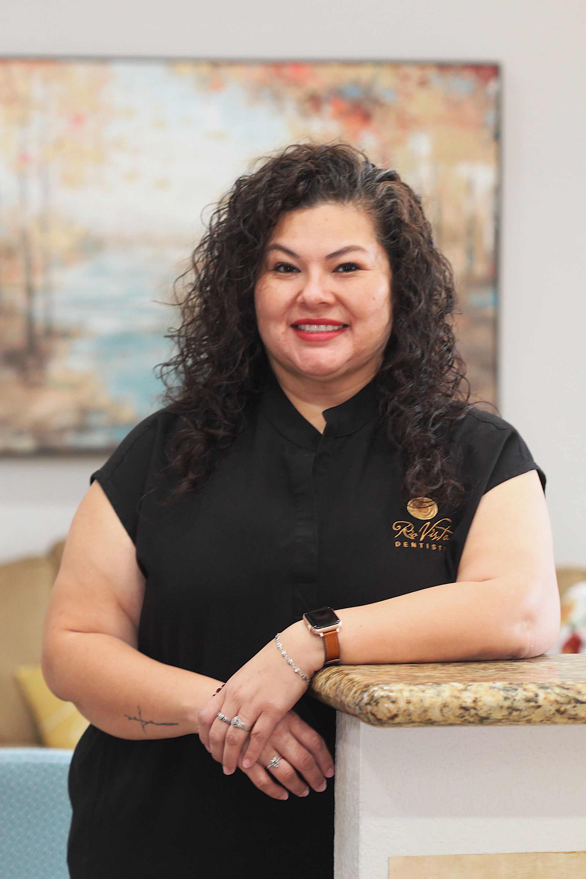 Woman in black top leaning on a counter, smiling in a softly lit office setting.