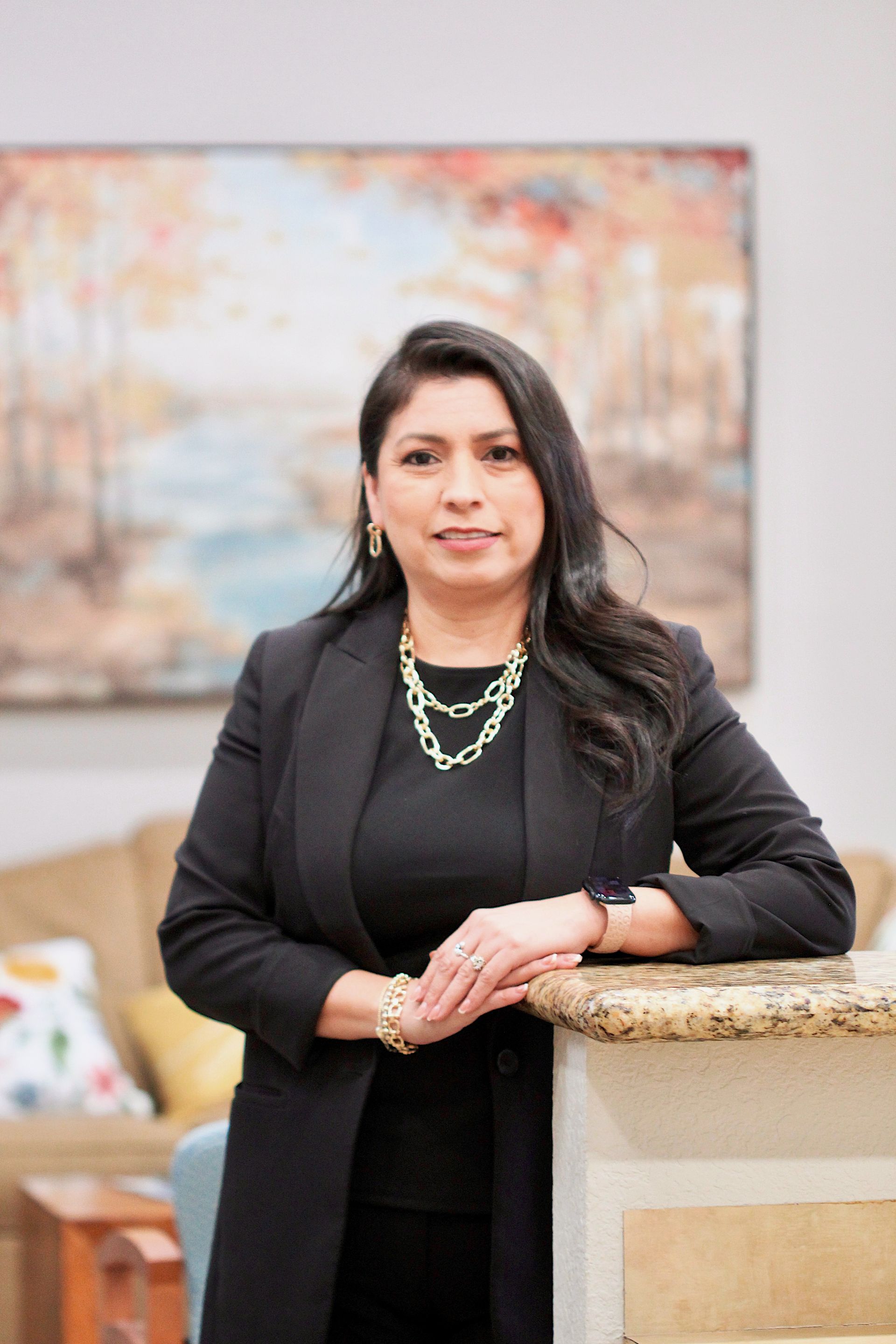 Woman in black standing by a stone counter, wearing layered jewelry, in a softly blurred indoor setting.