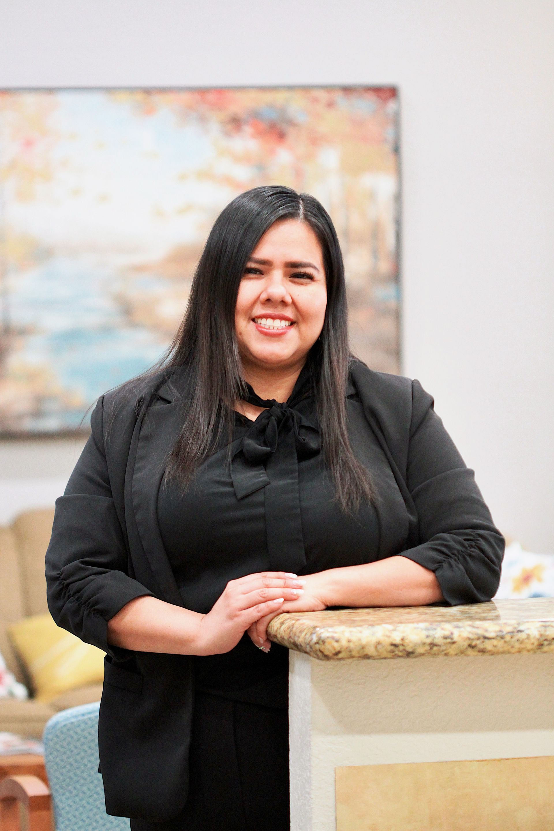 Smiling person in black blazer leaning on a stone counter indoors, with a colorful abstract painting behind.
