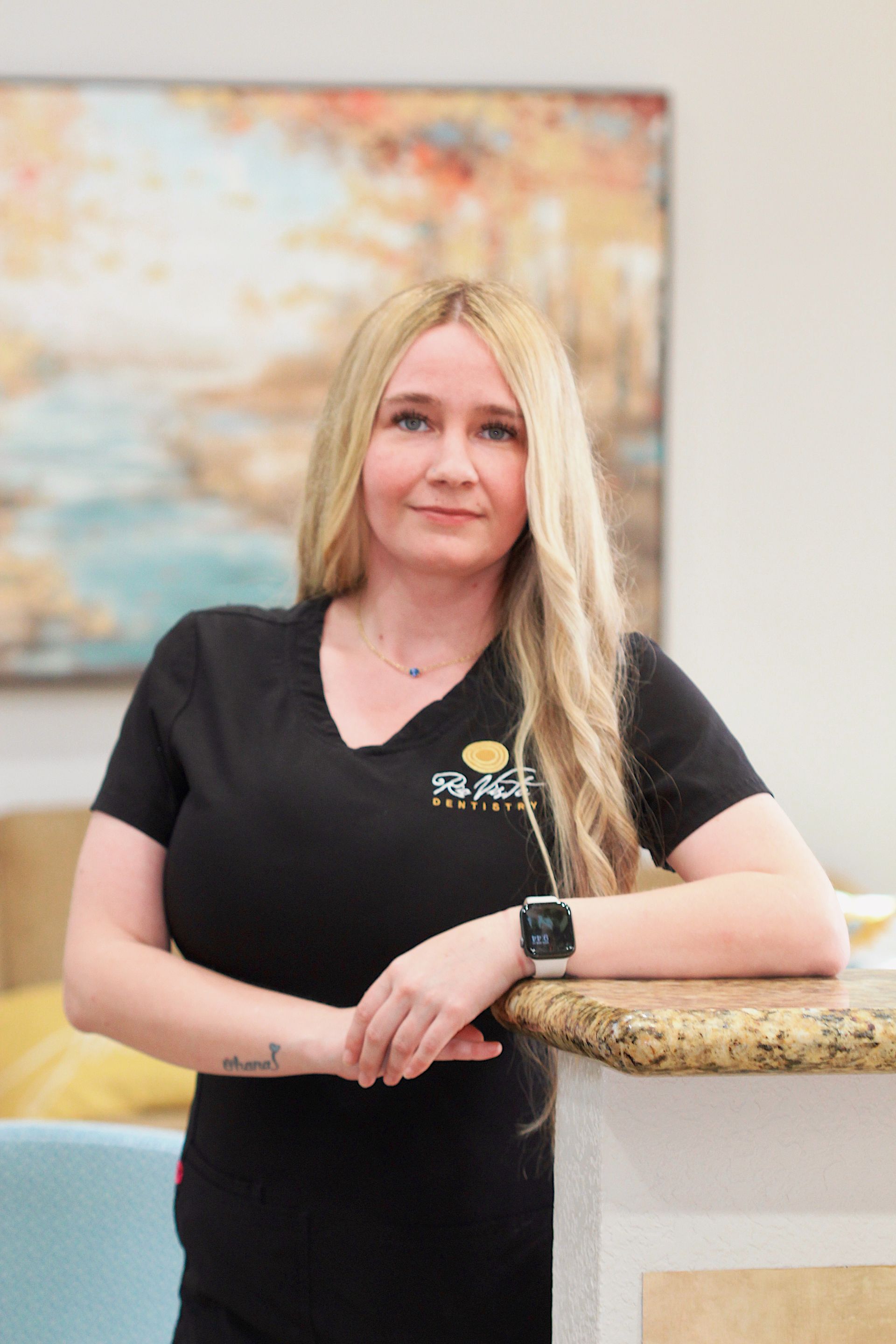 Woman leaning on a counter in a black shirt, smiling in a bright indoor setting.