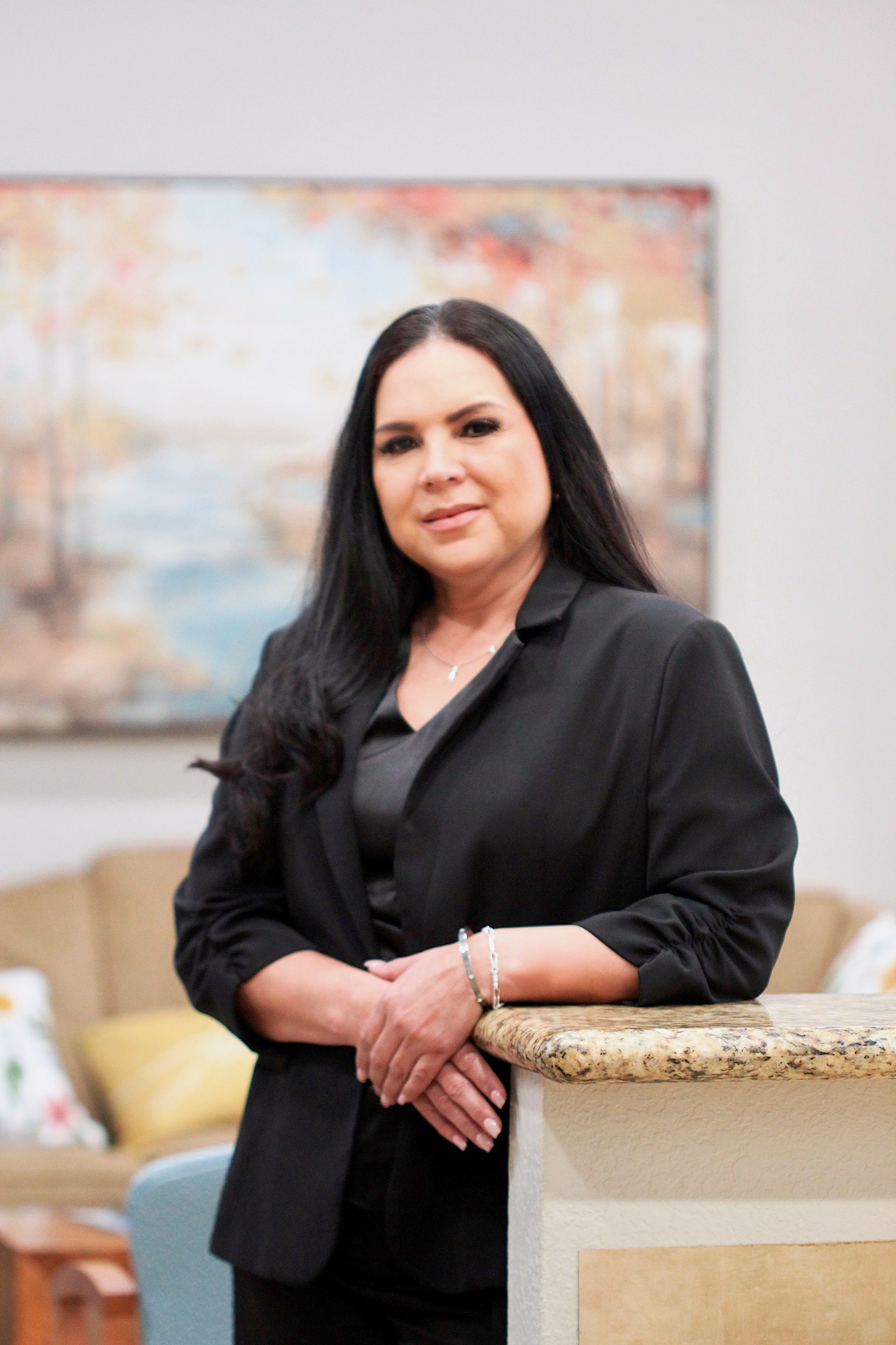 Woman in a black blazer standing indoors beside a stone counter, with a soft-focus painting behind her.