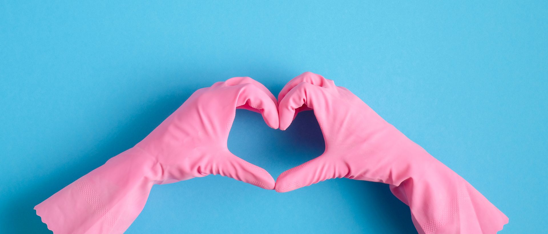 Two hands wearing pink rubber cleaning gloves shaped into a heart against a solid light blue background.