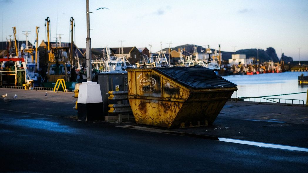 A weathered yellow dumpster on a dock; fishing boats and harbor in background.