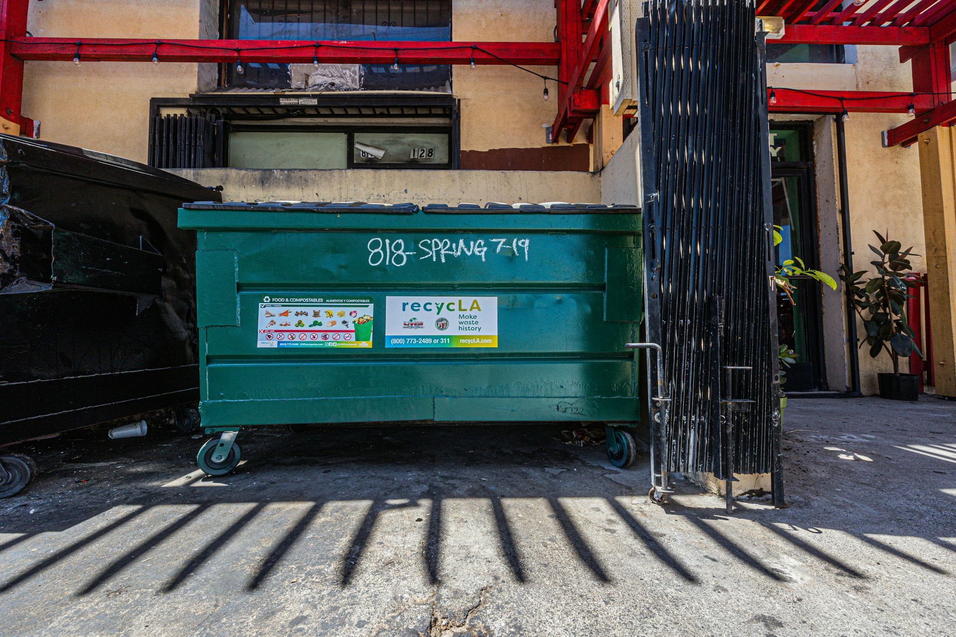 Green dumpster with blue and white labels, beside a black one and a metal gate. Shadows on concrete.