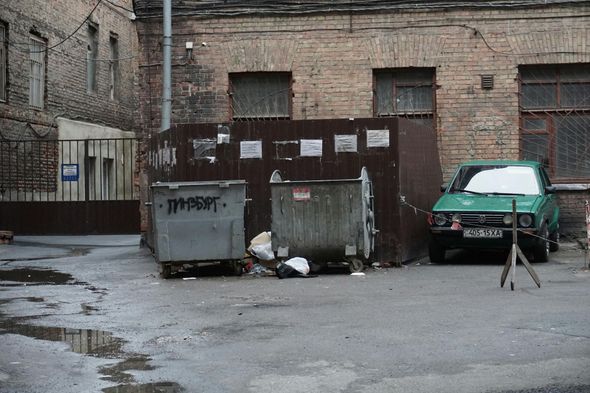 Green car parked near dumpsters in a brick-walled alley. Wet pavement, drab urban scene.