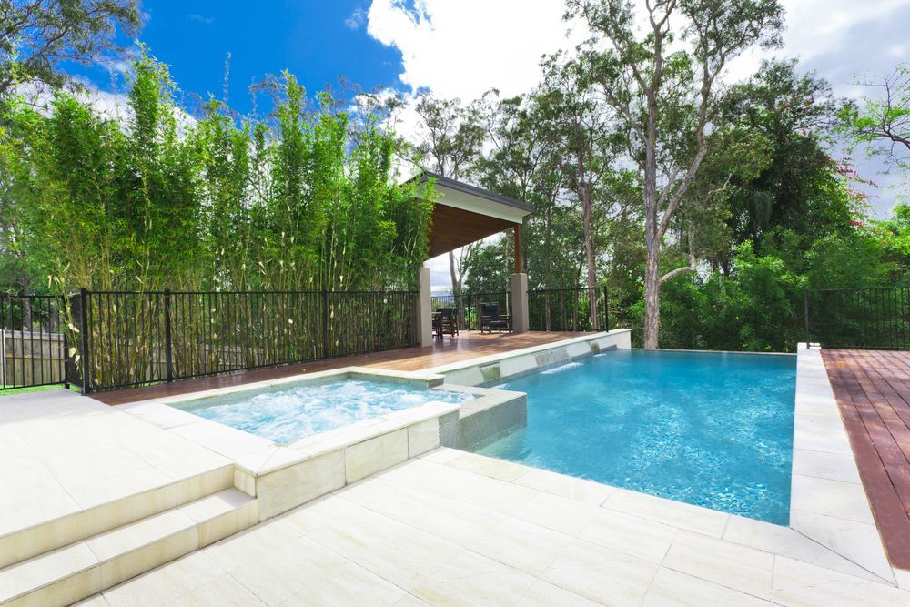 A backyard swimming pool and attached spa with light-coloured pavers, surrounded by bamboo and a covered patio area — Sand & Stone in Worongary, QLD