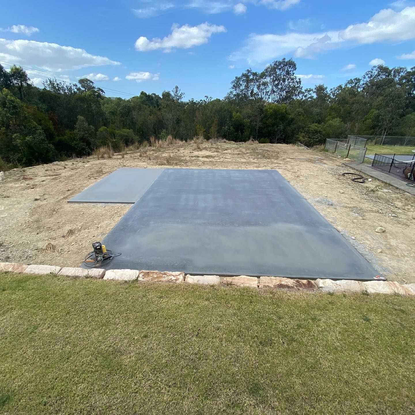 A freshly poured rectangular concrete slab sits on a cleared dirt plot in a sunny, grassy backyard landscape — Sand & Stone in Worongary, QLD