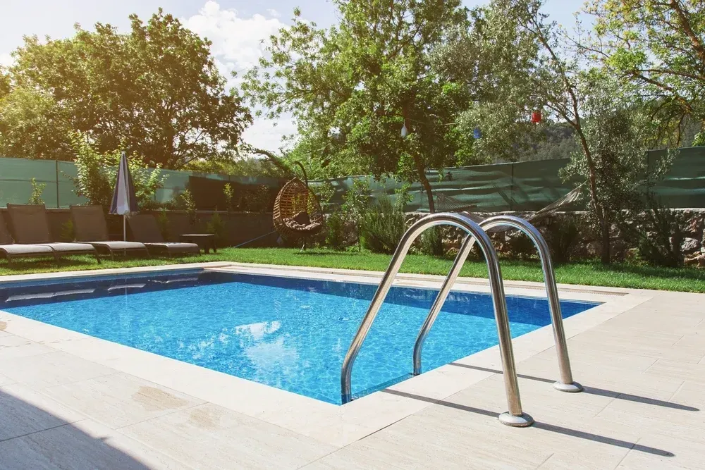 A bright blue rectangular swimming pool with metal ladder rails on a patio surrounded by trees and a green fence — Sand & Stone in Worongary, QLD