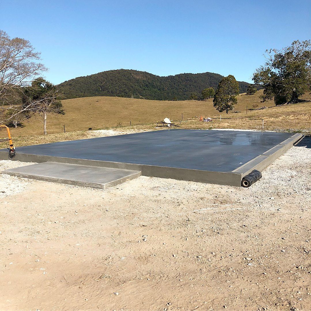 A freshly poured concrete slab foundation sits on gravel in a grassy, hilly rural landscape under a clear blue sky. — Sand & Stone in Worongary, QLD