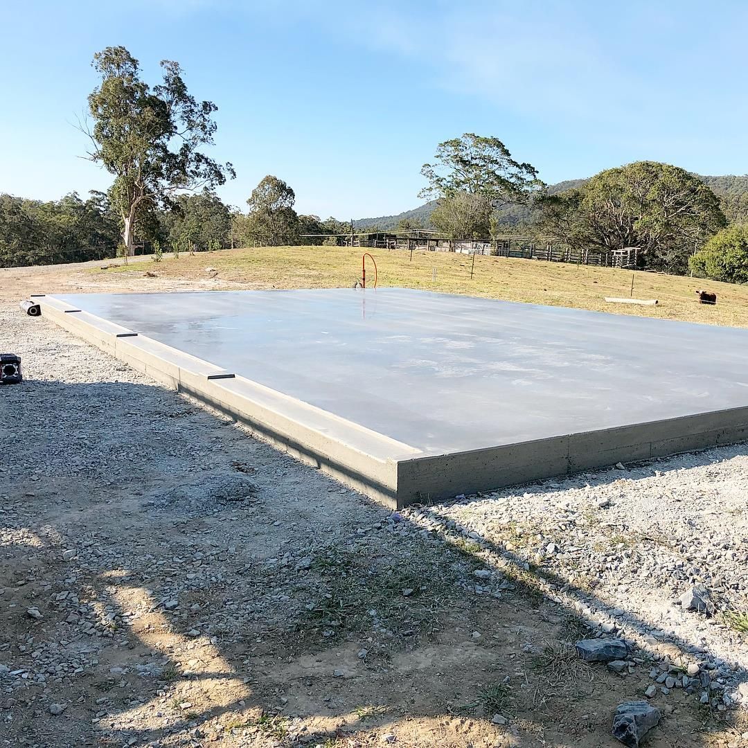 A freshly poured concrete slab foundation sits on a gravel base in a grassy, rural outdoor setting under a clear sky. — Sand & Stone in Worongary, QLD
