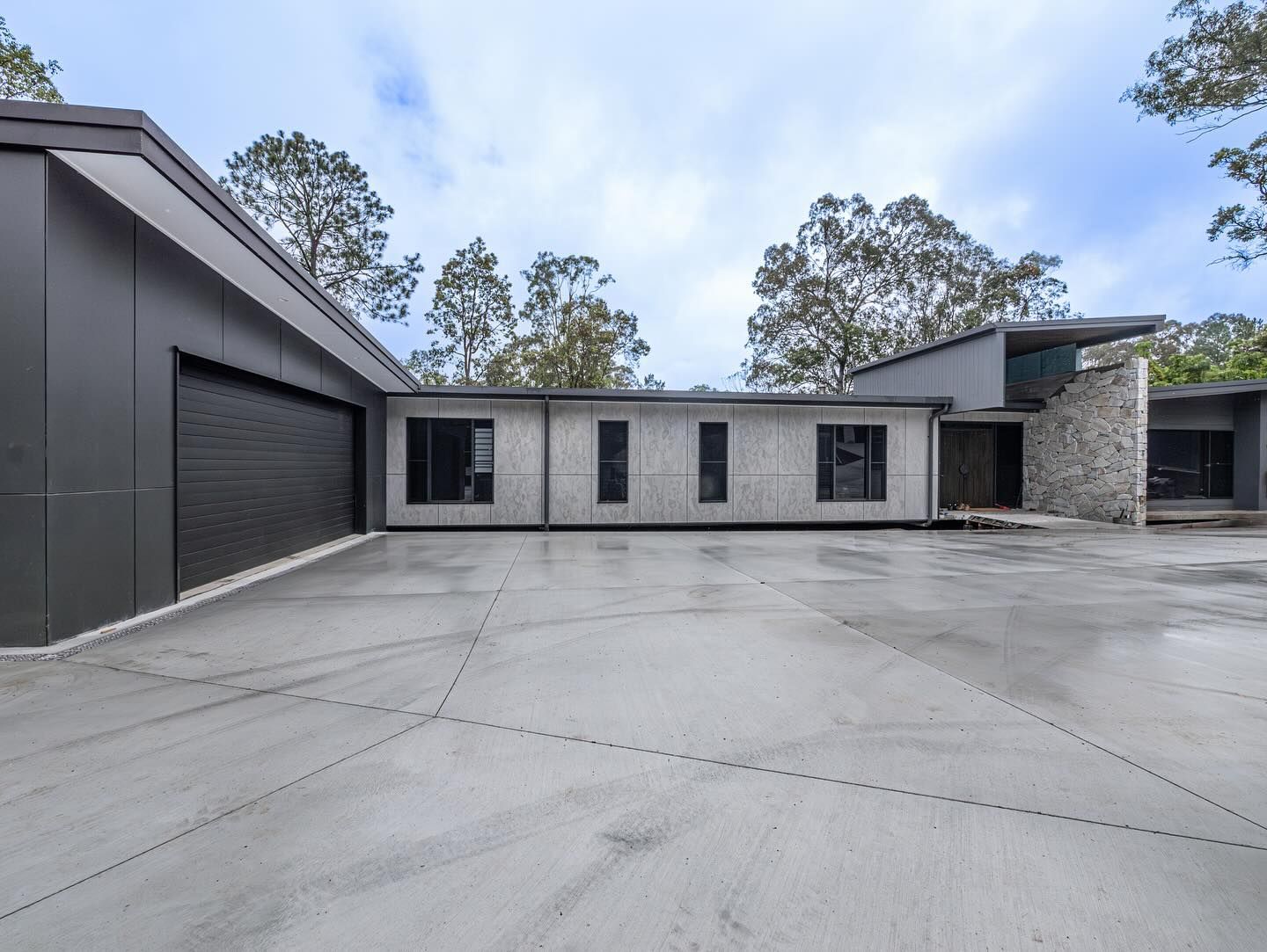 A modern home with a concrete driveway, dark gray garage, textured stone accent wall, and surrounding trees.