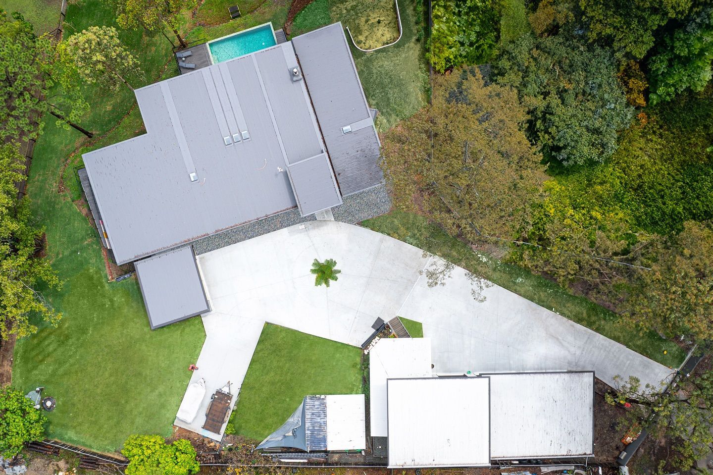 An aerial view of a modern home with a dark gray roof, a swimming pool, and a concrete driveway surrounded by trees — Sand & Stone in Worongary, QLD