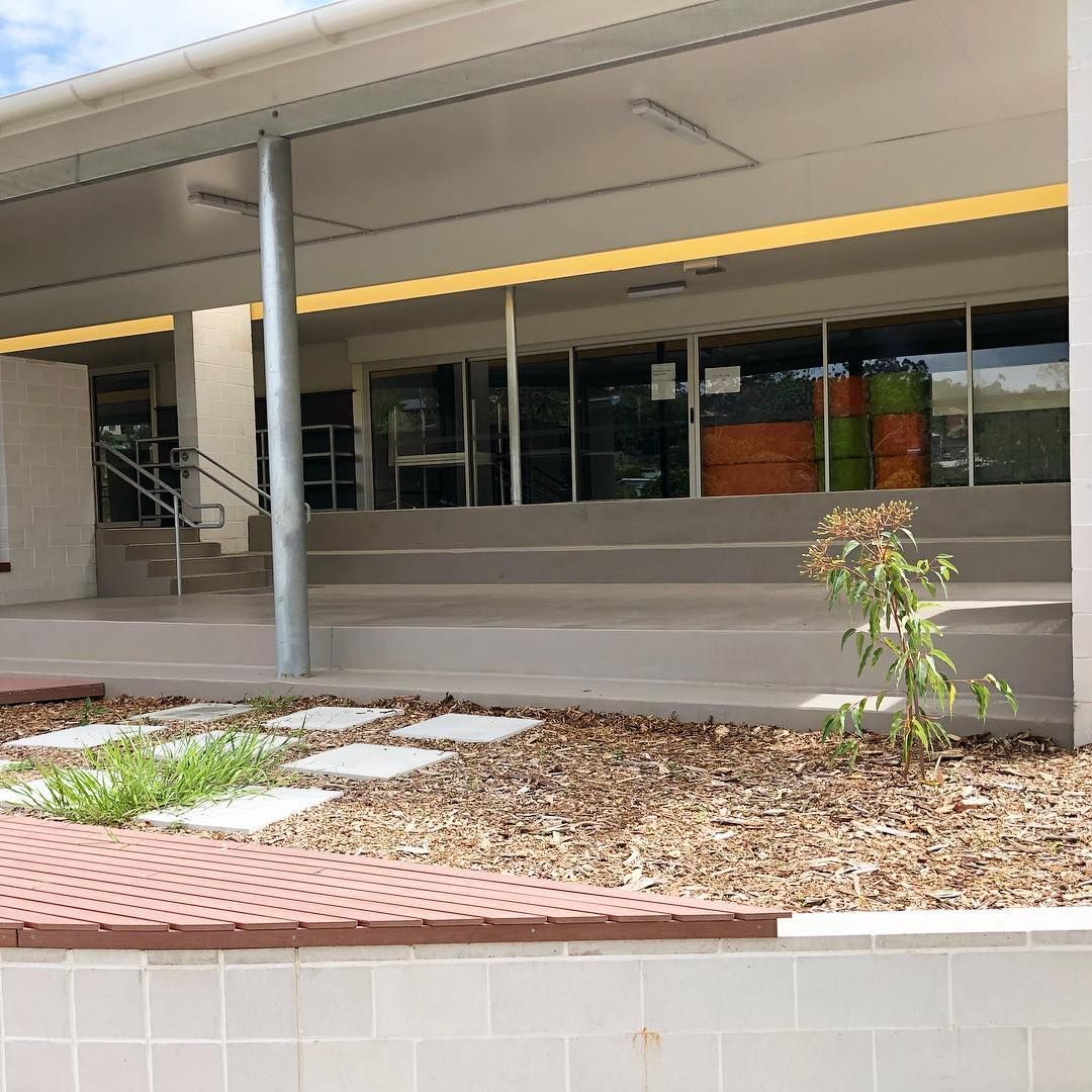 A building entrance with wide concrete stairs leading to glass doors, featuring a foreground garden bed and a metal pole— Sand & Stone in Worongary, QLD