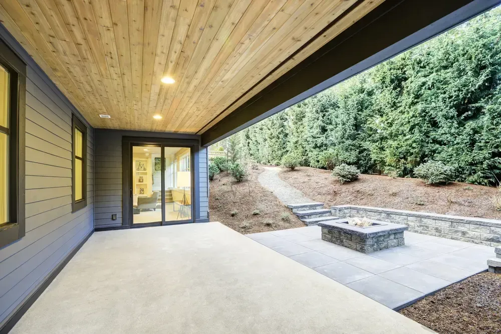 A covered concrete patio with light wood ceiling leads to an outdoor stone fire pit and a sloped path through the trees. — Sand & Stone in Tweed Heads, QLD