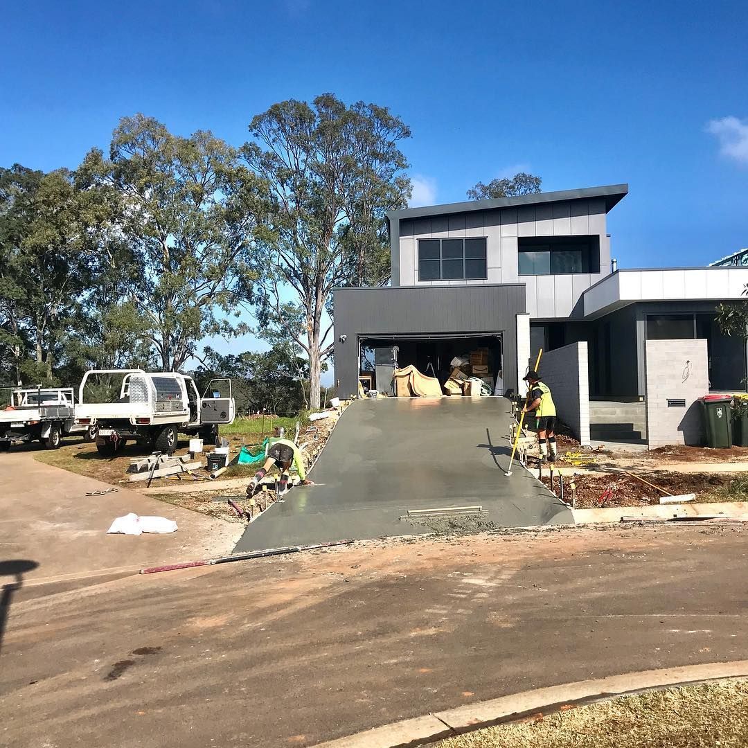 A modern gray two-story house under construction with a freshly poured concrete driveway and workers nearby. — Sand & Stone in Tweed Heads, QLD