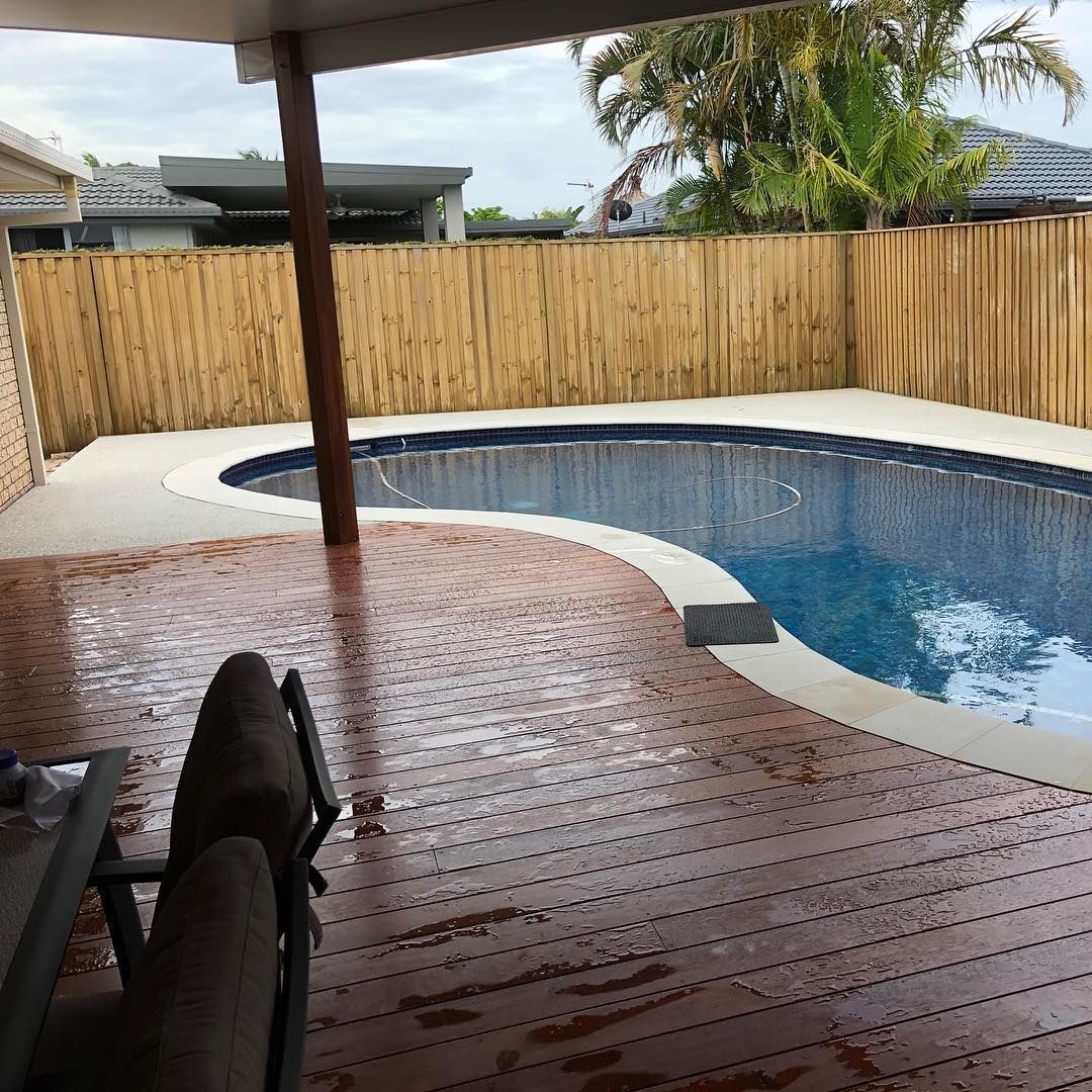 A wooden deck with a table and chairs overlooking a curved backyard pool surrounded by a light-coloured fence. — Sand & Stone in Worongary, QLD