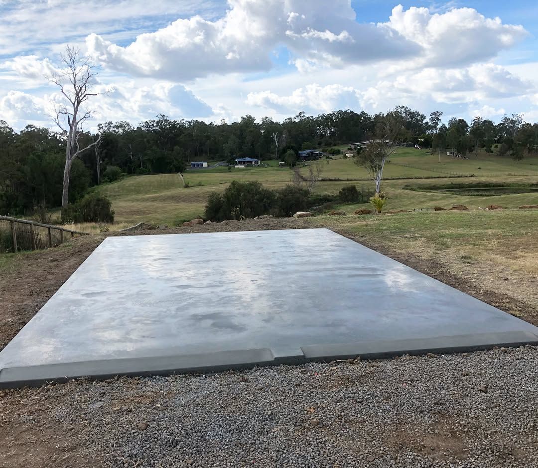 A freshly poured, smooth rectangular concrete slab set on a gravel base in a rural, grassy field under a cloudy sky. — Sand & Stone in Worongary, QLD