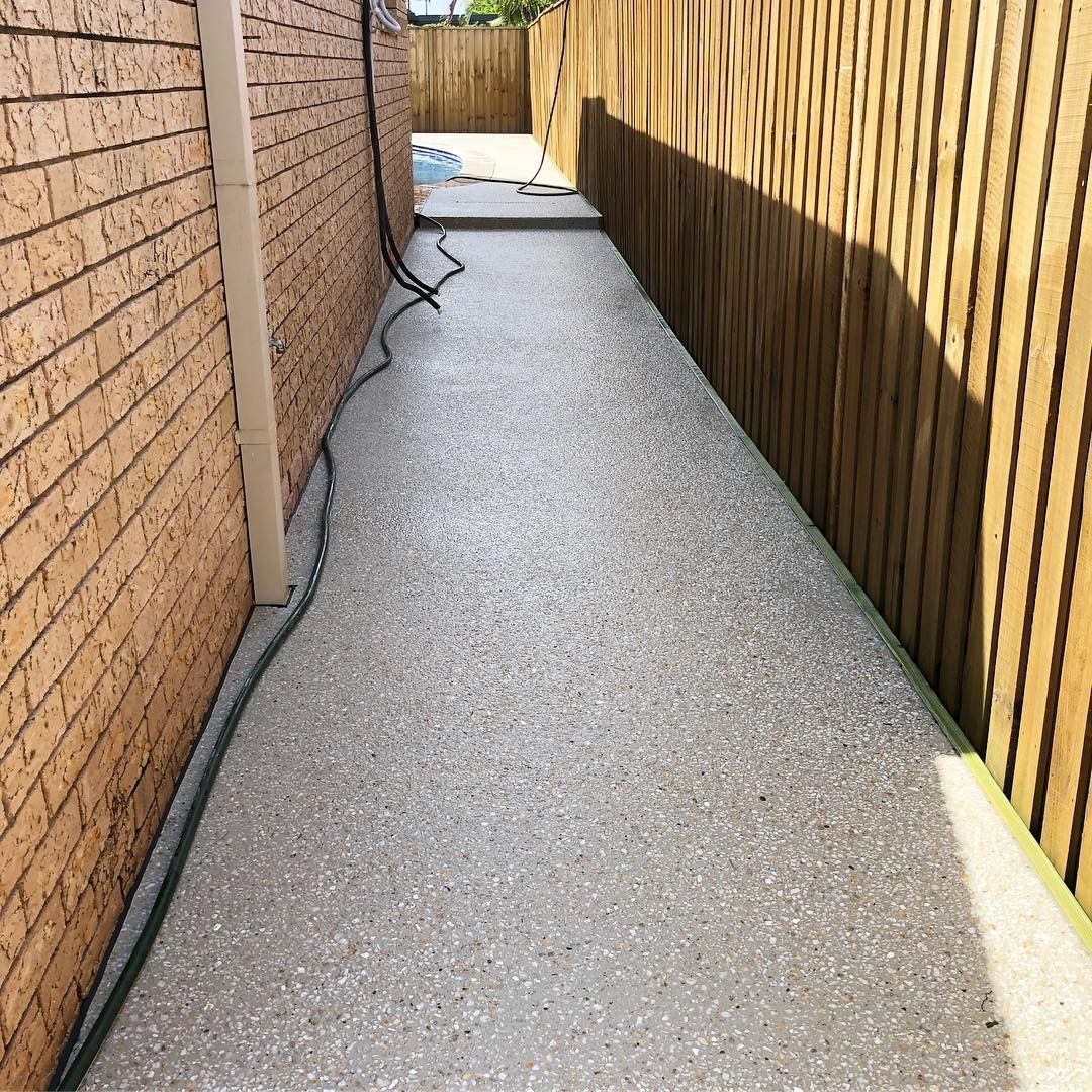 A narrow path with light gray speckled concrete flooring, bounded by a tan brick wall on the left and a wood fence on right. — Sand & Stone in Worongary, QLD