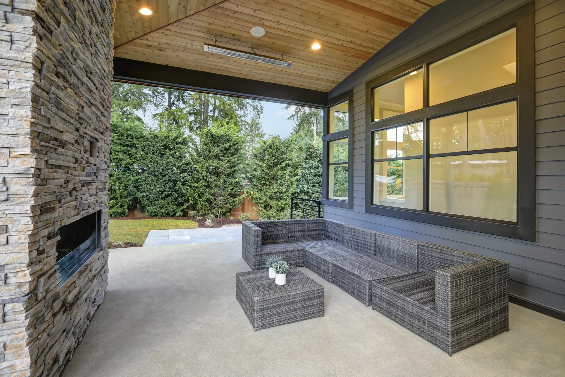 Modern patio with grey wicker sectional seating, a matching coffee table, stone fireplace, and large windows.