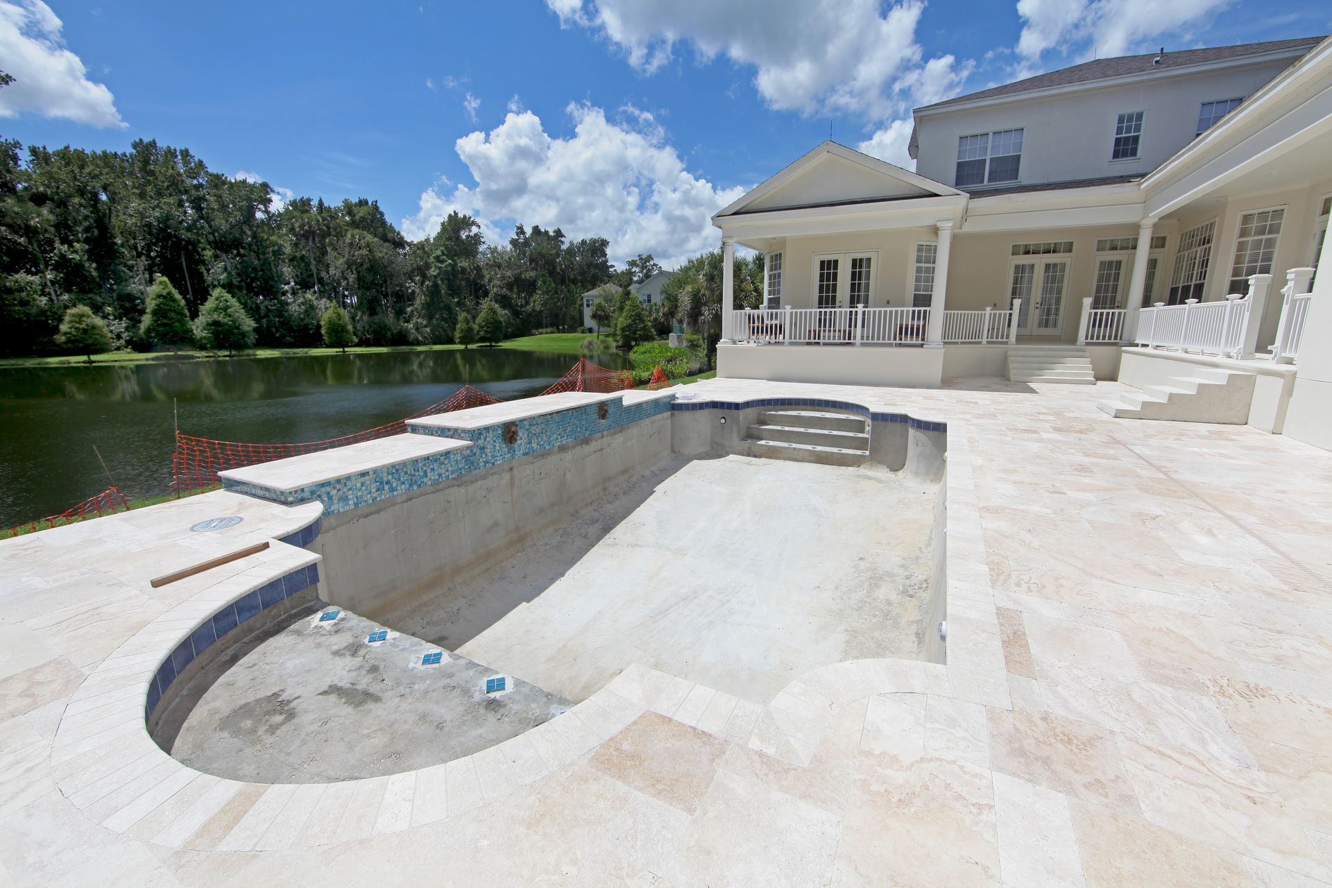A newly constructed, empty swimming pool with light stone tiling and blue accent tiles sits beside a pond and a house — Sand & Stone in Worongary, QLD