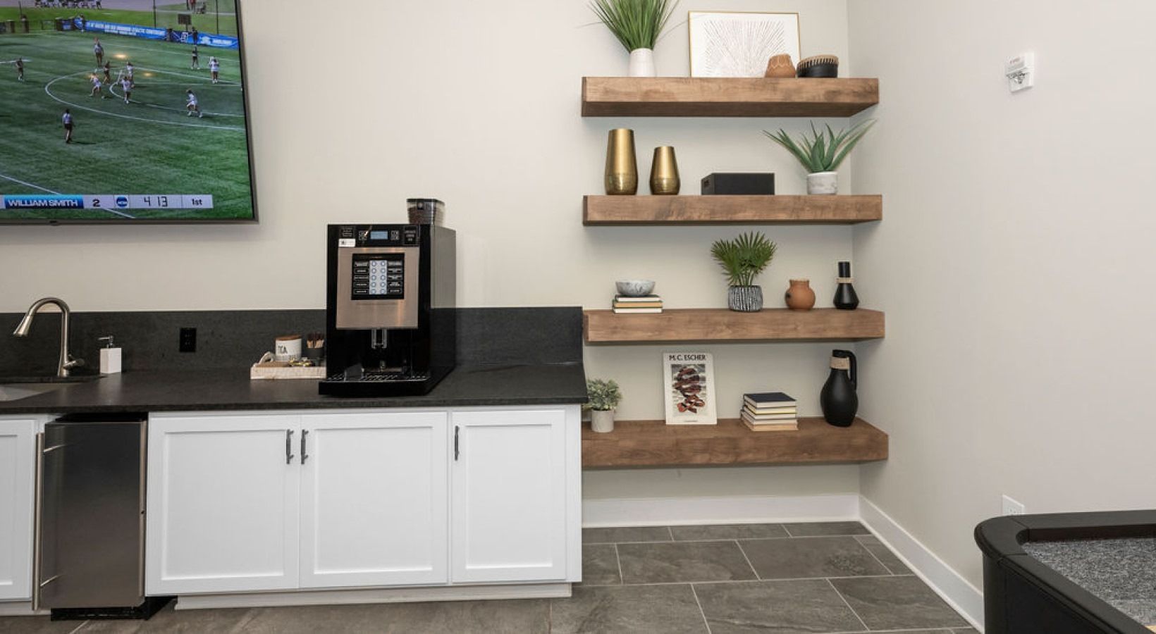 Coffee bar with white cabinets, dark countertop, shelves with plants, and a TV on the wall.  We invite you to visit 1001 Tyvola and take a tour of our pet-friendly, 3-bedroom apartments in Charlotte, NC.