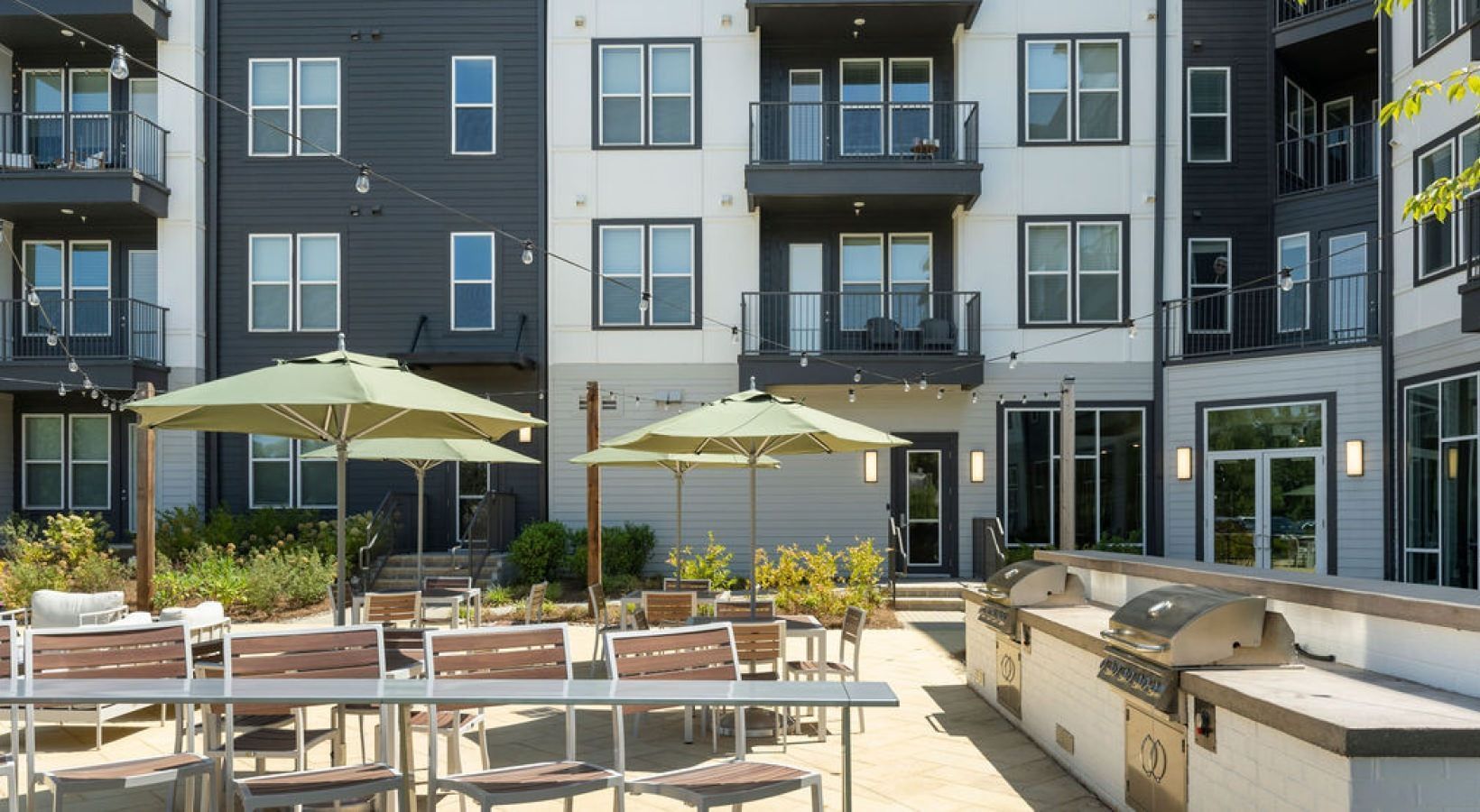 Outdoor patio with seating, umbrellas, and grills at an apartment complex with gray and white buildings.
