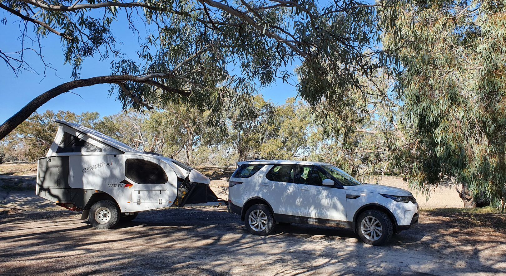 A white SUV is parked in a sunny, tree-lined clearing, towing a white teardrop-style camper trailer with its roof popped up.