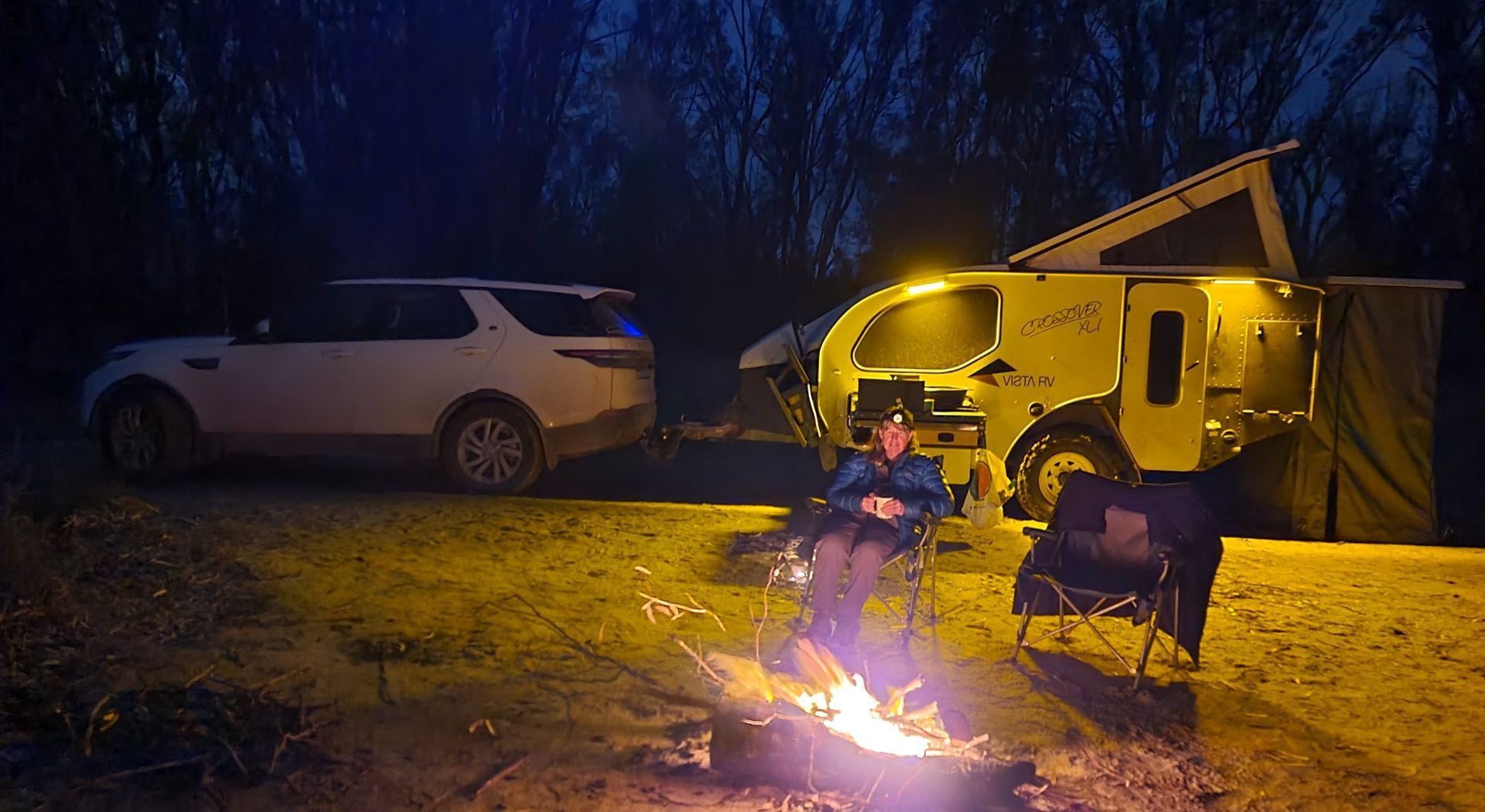 A person sits in a chair by a campfire at dusk, next to a white SUV towing a yellow pop-top camper trailer.