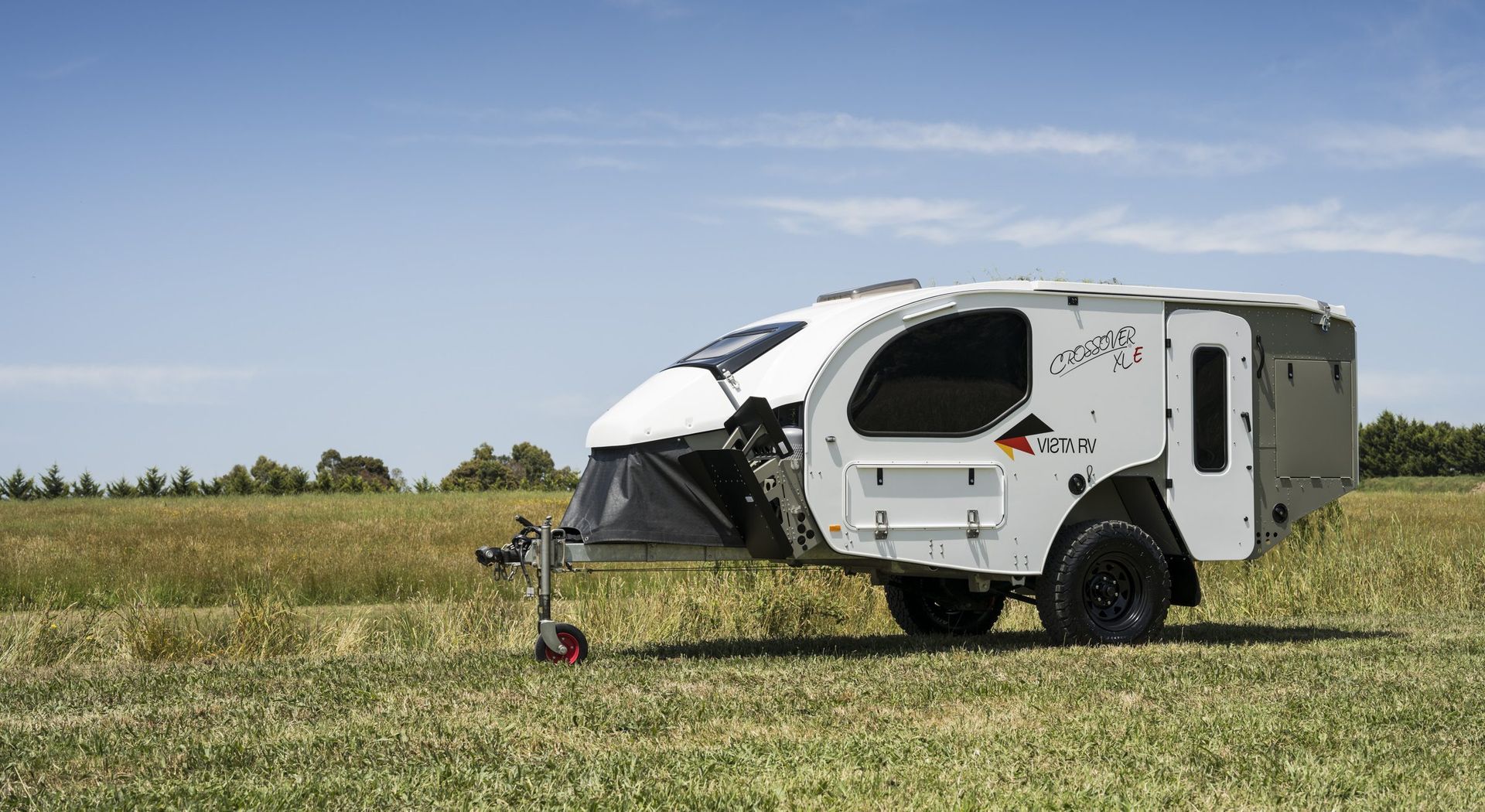 A white, compact off-road teardrop camping trailer sits in a grassy field under a bright blue sky.