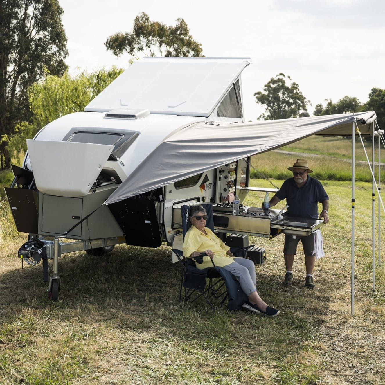 Two people camping by a pop-top caravan with an extended awning, outdoor kitchen, and folding chair in a grassy field.