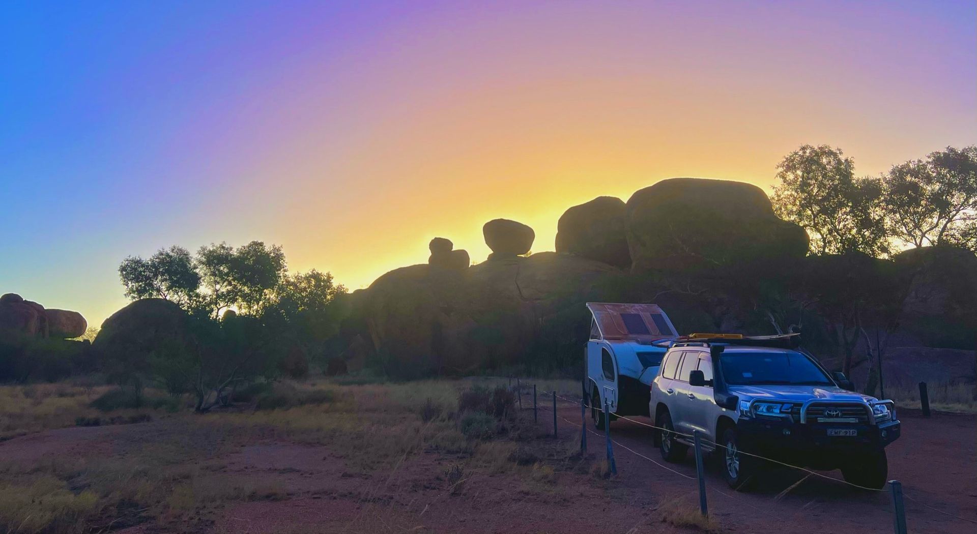 A white SUV towing a camper is parked in a desert landscape with large rock formations at sunset under a purple-blue sky.
