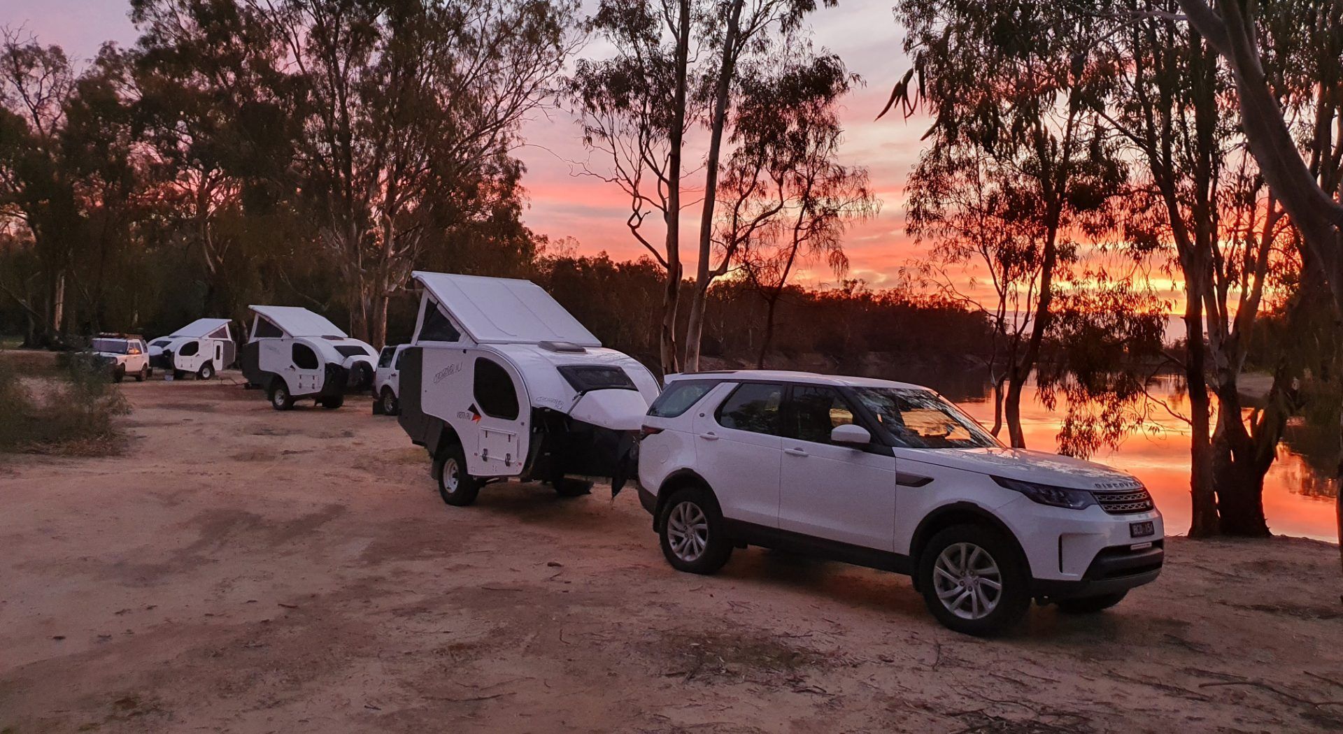 A white SUV tows a small white camper trailer at a campsite by a lake during a vibrant orange and pink sunset.