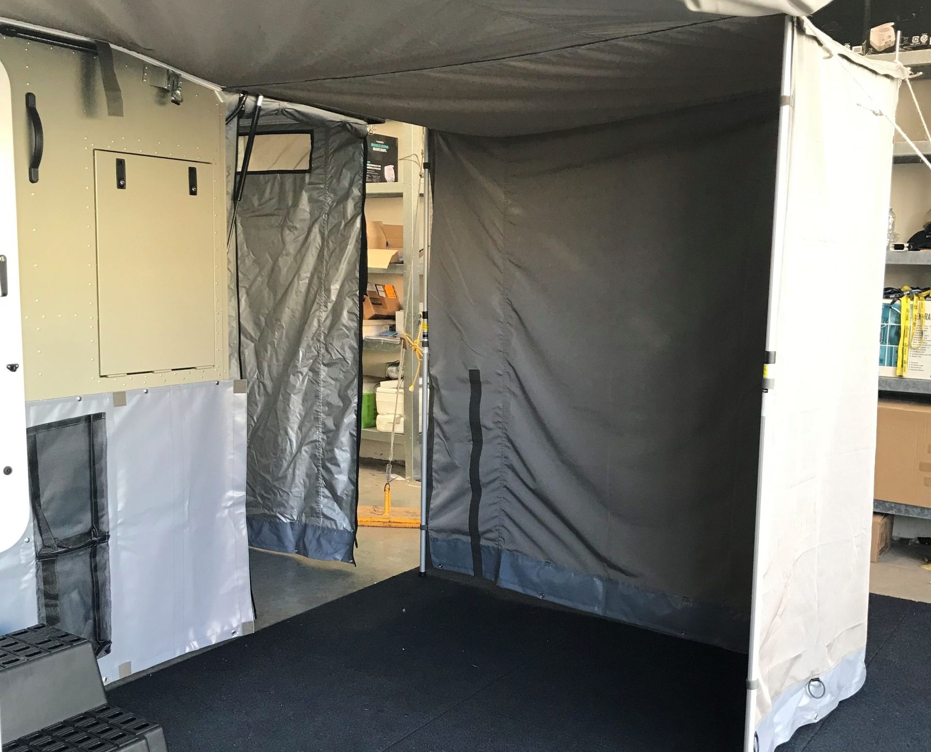 A tan privacy enclosure with a dark fabric interior and a black floor mat, set up beside a camper or vehicle.