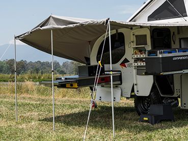 An off-road camper trailer parked in a field with an extended side awning and slide-out kitchen components visible.
