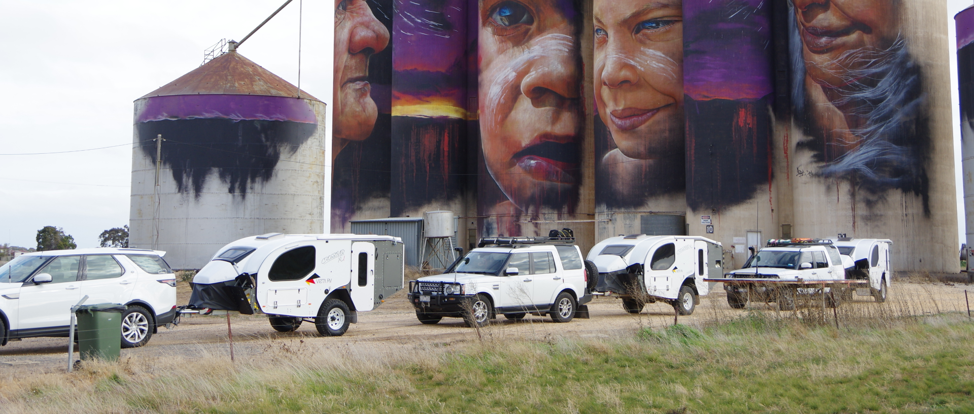 Several white SUVs and campers parked in front of tall grain silos featuring a large, vibrant portrait mural.