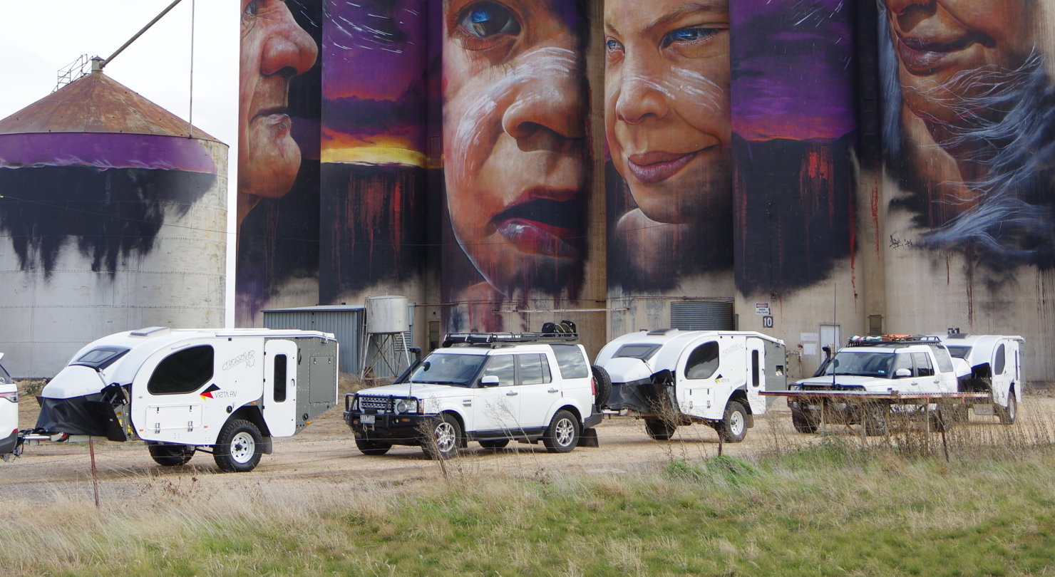 A line of white 4WD vehicles and camper trailers parked in front of tall silos decorated with a large, colorful mural.
