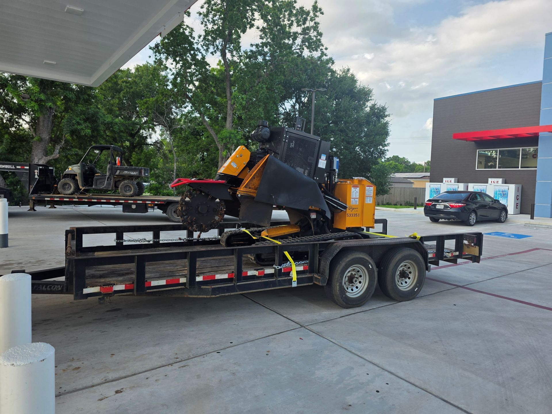 Construction equipment on a flatbed trailer at a gas station, with a yellow machine and black trailer.