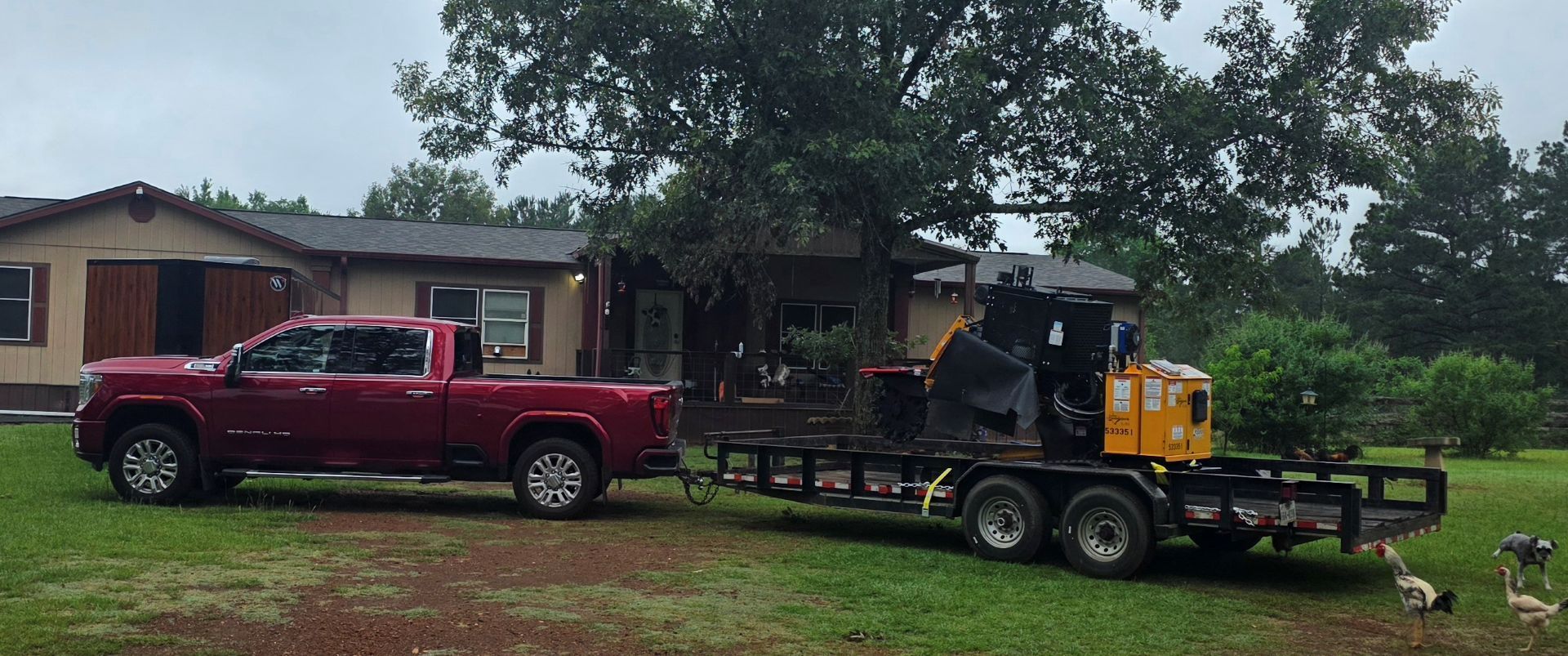 Red pickup truck and yellow trailer-mounted machine in a muddy yard beside a house under cloudy skies
